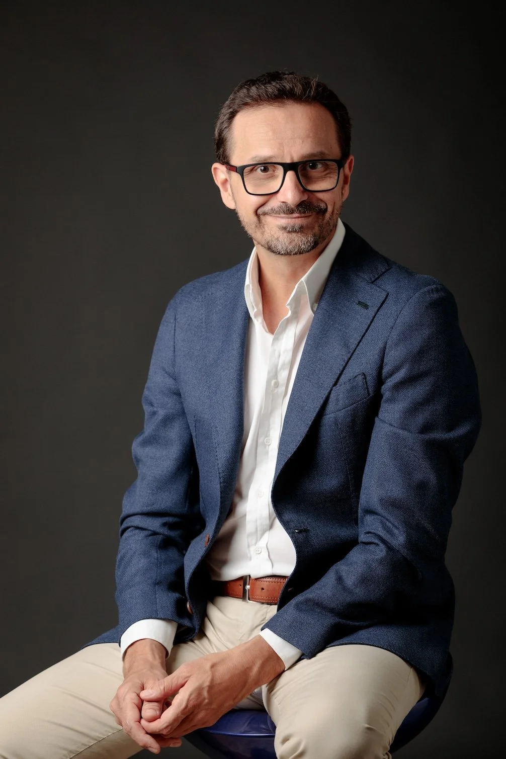 Uomo sorridente con occhiali sporchi, indossa giacca blu e camicia bianca, seduto su uno sgabello contro sfondo scuro.